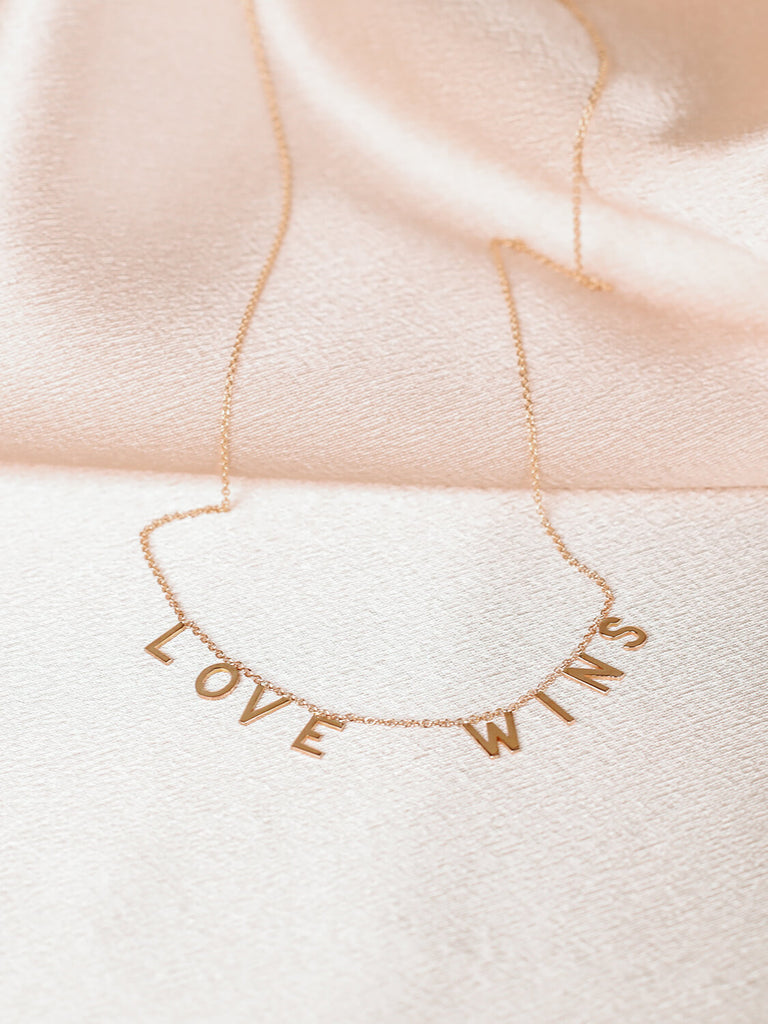 full view of "Love Wins" message collection necklace in yellow gold on pink satin background