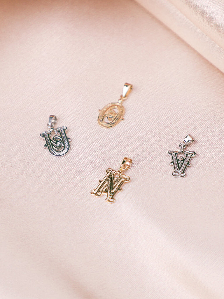 "U", "O", "N", and "V" gothic initial pendants in yellow and white gold laying on pink satin
