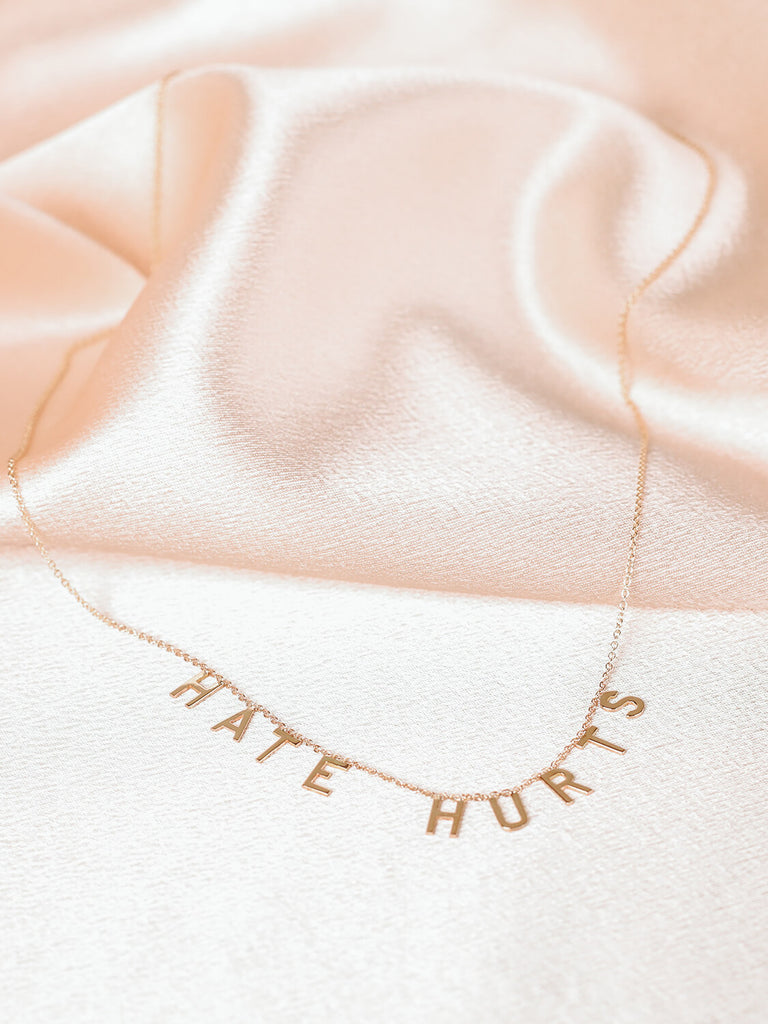 full view of "Hate Hurts" message necklace in yellow gold laying on top of pink satin