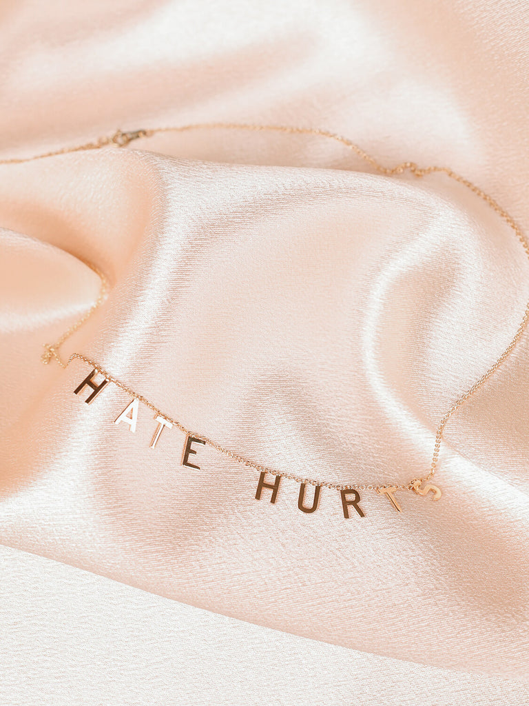 detail shot of "Hate Hurts" message necklace in yellow gold laying on top of pink satin