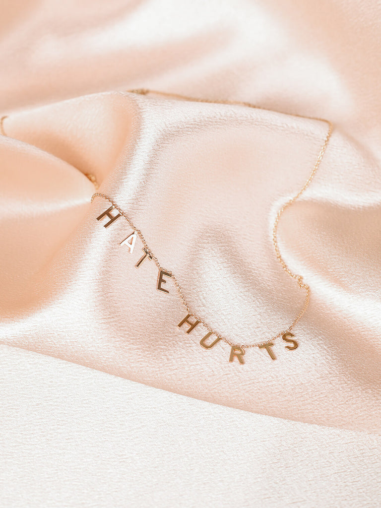 "Hate Hurts" message necklace in yellow gold laying on top of pink satin