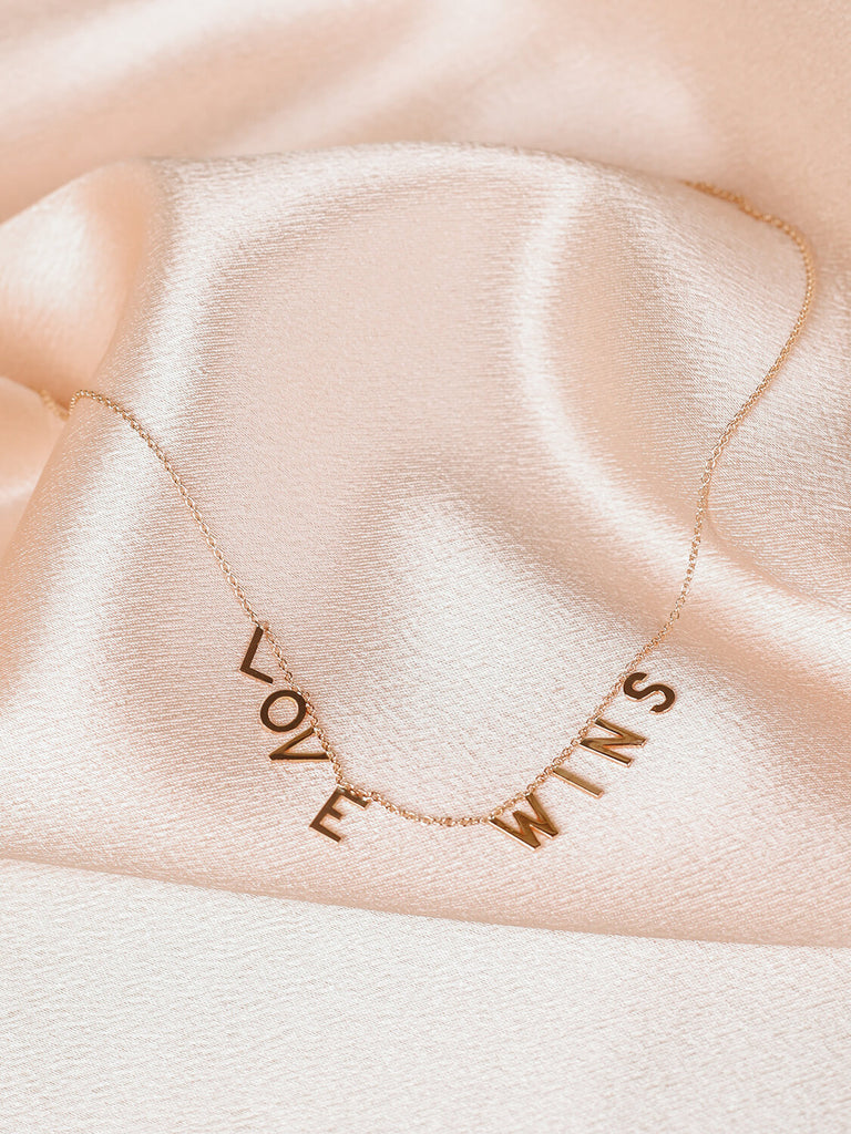 detail shot of "Love Wins" message collection necklace in yellow gold on pink satin background