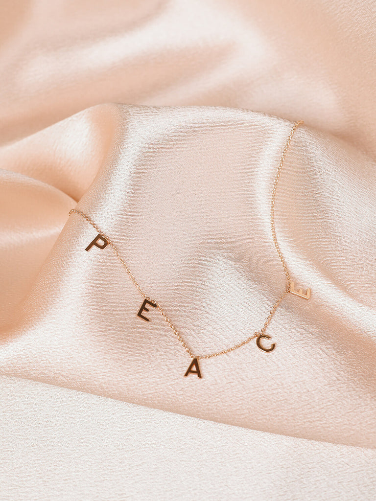 close up of "Peace" message necklace in yellow gold on pink satin fabric
